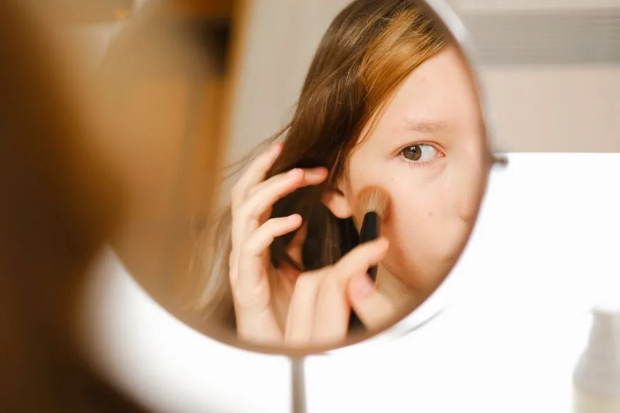 A young girl putting blush on her cheek while looking in a mirror.